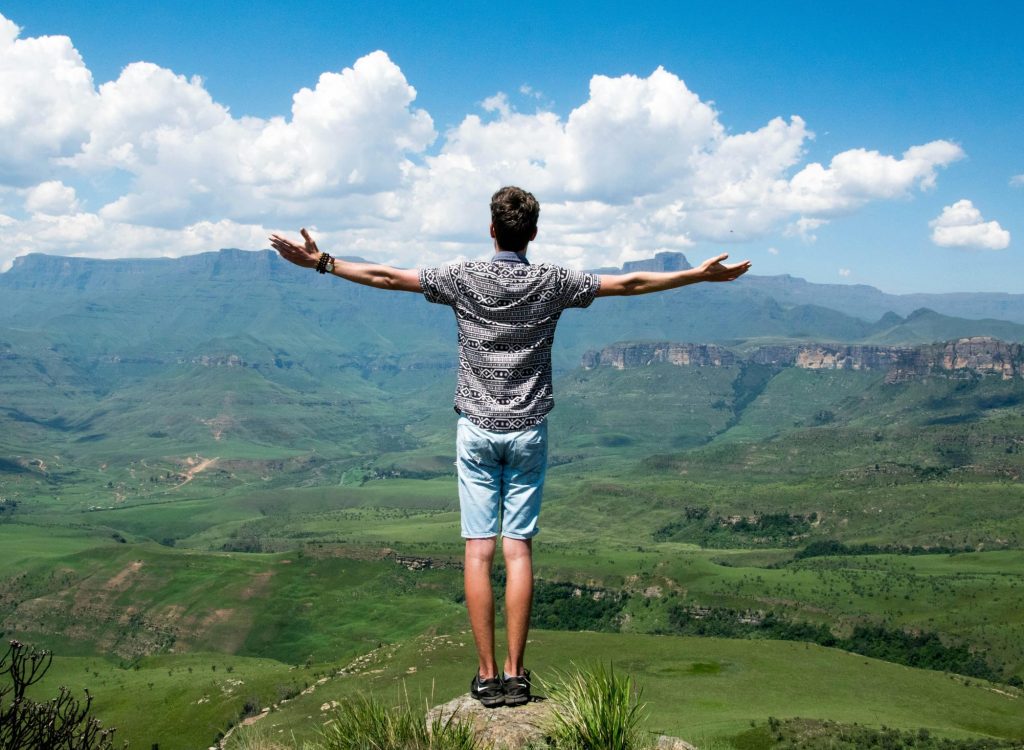 A man stands triumphantly with arms open, overlooking a scenic mountain range.